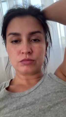 The media file features a close-up, head-and-shoulders shot of a woman with dark hair and fair skin, adorned with freckles. She is wearing a grey tank top and appears to be indoors, with sheer white curtains visible in the background. The lighting suggests it might be daytime. The woman is captured in a series of brief moments, with slight variations in her pose and expression. In some frames, her eyes are open and she looks directly at the camera with a neutral expression. In others, she closes her eyes or looks away, giving the impression of a candid, perhaps unposed, moment. Her right arm is raised, with her hand resting on her head or touching her hair in some shots. The overall mood is relaxed and natural. There is no visible text in the media file. The provided location context is Нижнеяркеево, Russia.