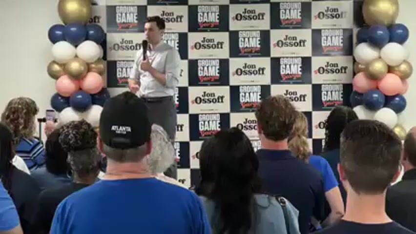 In an indoor setting in Decatur, United States, a man identified as Jon Ossoff is giving a speech to a seated audience. He stands at a podium or elevated platform, holding a microphone in his right hand and gesturing with his left. He is wearing a light-colored button-up shirt and dark trousers.

Behind him, a branded backdrop features a repeating pattern of "GROUND GAME Georgia" and "Jon Ossoff" logos in a checkered design. Two balloon pillars, consisting of blue, white, gold, and pink balloons, flank the backdrop on either side.

The audience, mostly seen from behind, is attentively facing the speaker. One individual in the foreground is wearing a black baseball cap with "ATLANTA UNITED" written on it. The event appears to be well-lit, suggesting either daytime or a brightly lit indoor venue.