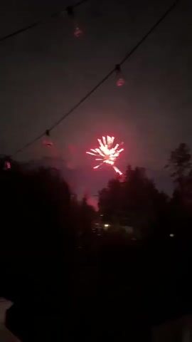 The video captures a nighttime fireworks display. In the foreground, silhouetted trees and what appear to be string lights are visible against the dark sky. A burst of white and yellow fireworks illuminates the sky with radiating sparks. As the display progresses, smaller, scattered fireworks and their trails of light are observed against the backdrop of a cloudy, dark sky. Some faint lights from houses or streetlights can be seen in the distance through the trees. The overall scene depicts an outdoor celebration with fireworks.