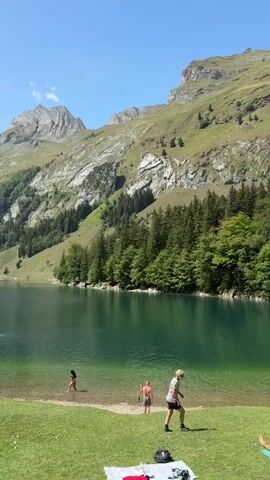 The video begins by showing a pristine, clear blue lake surrounded by lush green grass and dramatic, rocky mountains. Several people are visible by the lake, some swimming, while others are relaxing on the grass. The camera pans to show the vastness of the mountainous landscape, with dense forests covering the slopes. A pathway leads away from the lake, with a few people walking along it. The weather appears to be sunny with a clear blue sky. There are no signs of any buildings or artificial structures in the immediate vicinity.
