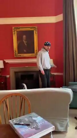 The video shows a man in a living room. He is wearing a gray hoodie, dark pants, and a hat with a blue and white pattern. He jumps onto the back of a gray couch and then immediately says "Oh my God, they're wicked," before then struggling to climb off the back of the couch. The room is decorated in shades of red. There is a painting hanging above the fireplace, as well as a table with a book and a pen on it. The camera is static and focused on the man and the couch.