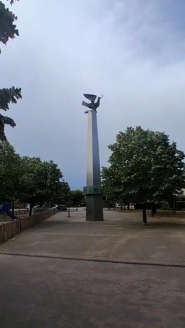 The video shows a tall, metallic obelisk with a bird sculpture on top, holding a branch. The obelisk has a square base with decorative panels. The scene appears to be an outdoor public space with trees and a cloudy sky.