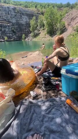The video shows a group of people at a quarry on a sunny day. In the foreground, two women are sitting on a beach towel. One woman is lying on her back in a transparent, glittery inflatable ring in the water, facing away from the camera. The other woman is sitting up, facing the water, wearing a black bikini top and black shorts. In the middle ground, a person is on a paddleboard in the water. In the background, a steep quarry wall with visible layers of rock rises above the turquoise water, with trees on top and along the sides. The ground is covered in gravel and dirt. Various items, including a cooler, shoes, bags, and drinks, are scattered on the beach.