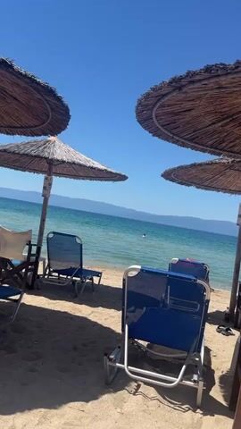 The video opens with a close-up of two straw umbrellas on a sandy beach. Beyond the umbrellas, the turquoise water of a lake or sea stretches towards a distant, hazy mountain range under a clear blue sky. Several blue beach chairs are visible on the sand, some unoccupied and some with another person sitting on them. A person is swimming in the water in the distance. The camera then pans slightly to the right, revealing more beach chairs and a partial view of a wooden structure on the right side of the frame. The video ends with a lingering shot of the beach, the water, and the umbrellas.