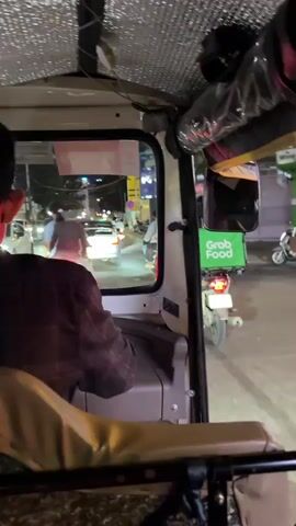 The video is taken from inside a tuk-tuk at night. A man is visible in the foreground, driving the tuk-tuk, facing away from the camera. Through the windshield, a street scene unfolds with traffic, including motorcycles and cars. One motorcycle in the foreground has a green "Grab Food" delivery box on the back. The environment appears to be a city street with buildings and streetlights. The video shows the tuk-tuk moving through the traffic.