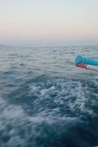This video shows a choppy sea from the perspective of a boat. In the foreground, white foam and waves churn behind the boat. The water is a deep blue, transitioning to a lighter blue in the distance. The sky above is a soft gradient of pale pink and gray, suggesting either dawn or dusk. In the far distance, faint, hazy mountains are visible on the horizon. On the right side of the frame, a portion of a blue and white object, possibly part of the boat's railing or a piece of equipment, is visible. The overall atmosphere is serene and vast, emphasizing the open water environment.