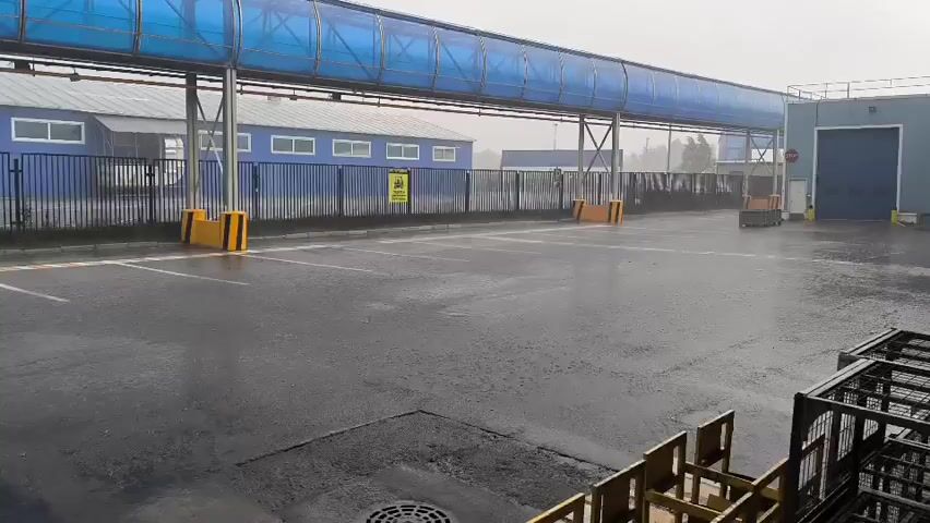 The video shows an outdoor industrial area on a rainy day. The ground is wet and reflects the light. A blue covered walkway stretches across the scene, supported by metal poles. In the background, there are blue buildings with windows, and a black fence. Yellow bollards are positioned at intervals along the fence line. A small yellow forklift is visible in the distance. To the right, part of a metal cage or structure is in the foreground. The overall atmosphere is grey and damp due to the rain.
