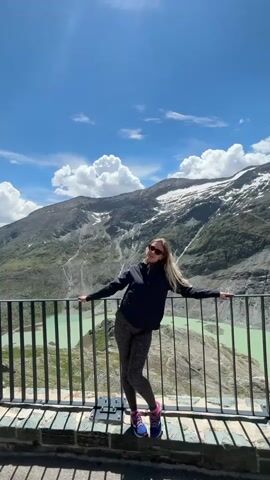 A young woman stands at a railing overlooking a turquoise lake and a mountain range under a bright blue sky with scattered white clouds. She is wearing a dark jacket, patterned leggings, sunglasses, and running shoes. Her arms are outstretched, leaning against the railing, and she is looking to her right. Her hair is blonde and appears to be blowing in the wind. The railing is made of metal and is supported by stone structures. The mountains in the background are rocky with patches of snow. The lake is wide and appears to be man-made, possibly a reservoir. The lighting suggests it is daytime.