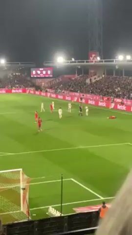 The video shows a soccer game at night. The score is 0-0. Players in red and white uniforms are visible on the green field. A player in a red uniform is lying on the ground. An official in a black and white uniform is standing with other players. The stadium is full of spectators. Advertising for Betclic and RE/MAX is visible on the sidelines. A soccer net is in the foreground of the shot.