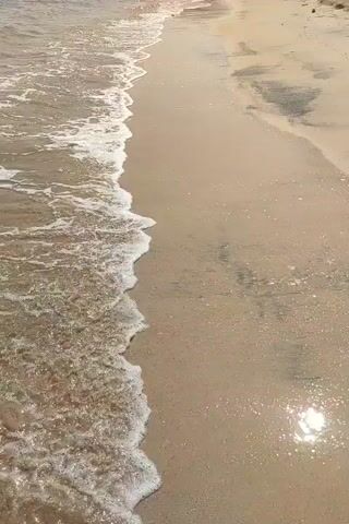 This is a video taken from a first-person perspective, looking down at a sandy beach and the edge of the ocean. The camera is moving forward, slowly approaching the water. Gentle waves with white foam are rolling onto the shore, leaving a thin layer of water on the sand. The sand itself appears wet and slightly darker where the water has receded. In the distance, along the shoreline, there are hints of vegetation and possibly some structures or umbrellas, suggesting a populated area. The overall lighting indicates it is daytime, with the sun glinting off the wet sand and water. There are no clear individuals visible in the immediate foreground, but there are some indistinct figures further up the beach in the distance. The soundscape is dominated by the gentle sound of waves.