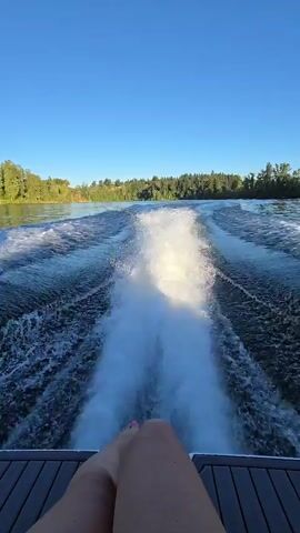 The video shows a first-person perspective from a boat moving on a body of water. A person's legs and feet are visible in the foreground, resting on the deck of the boat. The camera is facing backward, capturing the turbulent white wake created by the boat as it travels through the dark blue water. The wake stretches out behind the boat, fanning out and gradually dissipating. The surrounding environment consists of a clear blue sky above and a tree-lined shore in the distance, suggesting a natural, possibly recreational setting. The lighting indicates it is daytime, with the sun casting a bright light on the water and the wake.