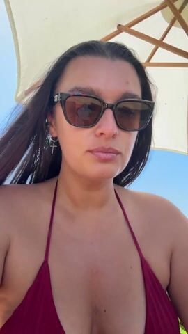 A young woman wearing sunglasses and a burgundy bikini bites into a sandwich. She is outside, presumably at the beach as the sky is blue and an umbrella is overhead. She chews the sandwich, looking directly at the camera. The audio appears to contain music.