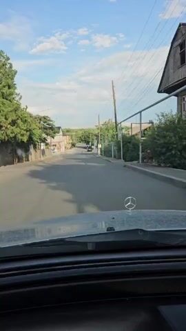 The video shows a forward-facing view from inside a vehicle driving down a paved road lined with trees, houses, and utility poles. The sky is blue with scattered clouds, suggesting daytime. A Mercedes-Benz hood ornament is visible on the car's hood. The road appears to be in a residential area, with a few other cars visible in the distance.