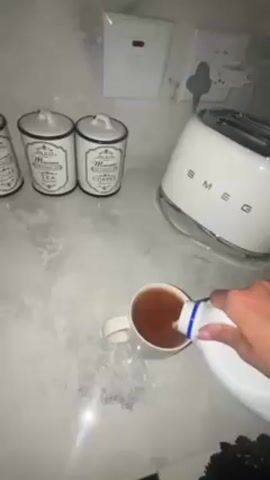 Here's a description of the video:

A person is adding milk to a mug of tea. The mug is on a light-colored countertop. The person's hand holds a white container, presumably filled with milk, as they pour it into the mug. 

In the background, there's a white Smeg toaster and several canisters labeled "Tea," "Coffee," and "Sugar." Also visible are a light switch and an electrical outlet.