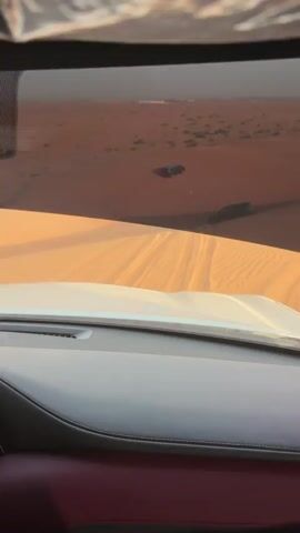 This video shows the view from inside a car driving on a sand dune. The car is white, with a black dashboard and red interior. The windshield sunshade is down, covering part of the frame. Through the windshield, there are other cars driving on the dunes. The sand is orange. The sky is light, but there is some fog or haze in the distance. The cars are creating tracks in the sand.