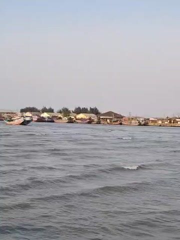 The video shows a body of water with small waves. In the background, there is a shoreline with numerous small boats docked. Some buildings and trees are visible on the shore. The sky is clear and pale. The camera is positioned low, likely from another boat, looking towards the shore. There are no people visible in the video. The overall scene depicts a harbor or a coastal village with fishing boats.