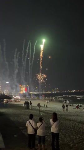 People are watching fireworks at night on a beach. There are buildings in the background, and people are walking around on the sand. Three people in the foreground are facing the fireworks.