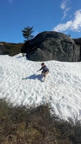 The video depicts a person throwing a snowball on a snow-covered landscape. The person, wearing a dark jacket and patterned trousers, is positioned on a snow-covered slope, reaching down to form a snowball, then stands up to throw it before crouching down again. The ground is covered in snow, with visible footprints. Behind the person is a large dark rock formation and a sparse evergreen tree. The foreground shows patches of green and brown vegetation. The sky is bright blue with scattered clouds. The camera briefly pans to the left, revealing more of the snowy landscape before returning to focus on the person.