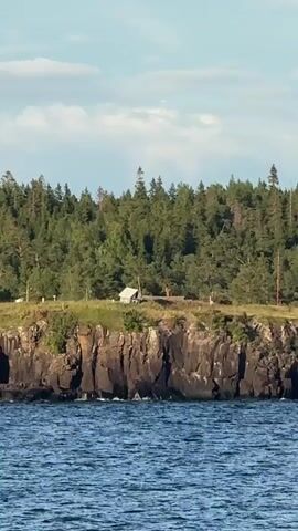 The video shows a wide shot of a body of water with a rocky, tree-lined coastline in the background. There are a few small structures, possibly cabins or tents, visible on the grassy clifftop. The water is choppy with small waves. The sky is partly cloudy with a clear blue expanse. The camera appears to be on a boat, as the railing and some seating are visible in the foreground. The overall scene is peaceful and natural, likely taken on a summer day.