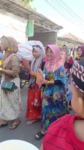 The video opens with a group of young people, mostly women, dressed in colorful clothing and hijabs, walking down a street. They are holding what appear to be decorative objects or props. One woman in the foreground is wearing a pink hijab and a blue and green patterned dress, and she is looking at her phone. Another woman next to her is wearing a red and blue patterned dress and a light blue hijab. A third woman in a blue dress and maroon hijab is laughing. In the background, a man is pushing a bicycle with a large sack on the back. A motorcycle with a rider wearing a helmet is also seen. The scene appears to be outdoors, on a paved street, with buildings and trees visible. The overall atmosphere seems festive or celebratory.
