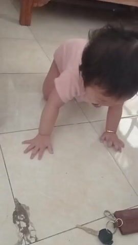 A baby in a pink onesie is crawling on a tiled floor. There are keys and a key fob on the floor in front of the baby. The baby looks at the camera with a neutral expression.