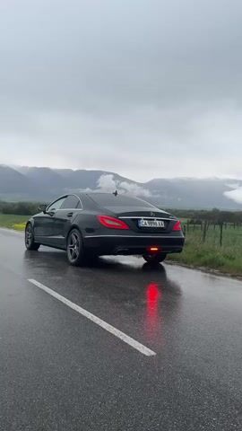 A black Mercedes-Benz CLS (likely a W218 model) is parked on the wet asphalt shoulder of a road, viewed from a rear-three-quarter angle. Its brake lights are illuminated, casting a red glow on the wet surface. The car displays a Bulgarian license plate, "CA 9898 XA". To the right of the road, there's a grassy verge with yellow wildflowers and a wooden fence. In the background, majestic mountains rise, their peaks partially obscured by dense clouds or mist, suggesting an overcast and possibly damp day. The sky above is uniformly grey and cloudy. The road itself is paved and features a clear white center line. The overall scene suggests a quiet, rural setting after recent rainfall, with the car sitting still amidst a picturesque, albeit moody, landscape.