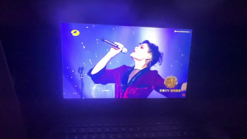 A TV screen displays a performance by a singer on a brightly lit stage. The singer is wearing a red outfit and is holding a microphone, singing the words "Purple rain, Purple rain" as shown on screen in both English and Chinese. The stage is illuminated with bright white and blue lights, and there is an audience visible in the background. The screen also displays the logo for "Singer", presumably the name of the show. The TV screen is being filmed in a dark room, with a laptop keyboard visible at the bottom of the frame.