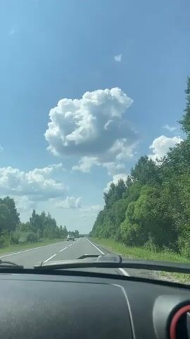This is a first-person perspective video recorded from inside a vehicle traveling on a paved road. The road has two lanes with a white dashed line separating them. The vehicle appears to be moving at a moderate speed. To the right of the road, there is a dense, green forest with tall trees. To the left of the road, there are scattered trees and some green vegetation. The sky is bright blue with several large, fluffy white clouds. One particularly prominent cloud is centered in the frame. In the distance, another white vehicle is visible on the road, traveling in the same direction. The dashboard of the vehicle from which the video is being recorded is partially visible in the foreground. The scene suggests a sunny day, possibly in a rural or semi-rural area.