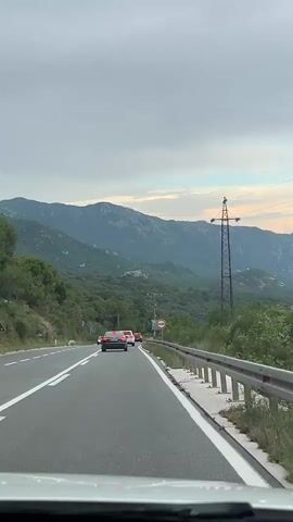 This video captures a scenic drive through a mountainous landscape in Stanišići, Montenegro. The camera is positioned inside a vehicle, looking forward and to the side. The time of day appears to be late afternoon or early evening, as suggested by the soft, diffused light and the muted colors of the sky. The weather seems overcast with patches of blue sky visible, indicating a dynamic sky.

The primary subjects are the road and the surrounding natural environment. A highway with white lane markings stretches ahead, with a few cars visible in the distance. A guardrail runs along the right side of the road, separating it from a steep, vegetated slope. Tall, dark electricity pylons with overhead wires are prominent features of the landscape, a common sight in many rural or mountainous areas. The mountains themselves are covered in lush green vegetation, rising majestically in the background.

As the vehicle continues, the view opens up to reveal a breathtaking vista of the Adriatic Sea in the distance. The water is a calm, deep blue, meeting a hazy horizon. On the coastline, a town or village is visible, with buildings clustered together and a few structures that suggest a harbor or marina. The greenery from the hillsides extends down towards the sea, creating a beautiful contrast between land and water.

There are no people explicitly visible within the frame, except for a reflection of a ring on a finger in the side mirror. The overall mood is one of peaceful travel and appreciation for the natural beauty of the Montenegrin coast. A traffic sign with a red circle and a white horizontal bar (indicating a prohibition or restriction) is visible on the side of the road, but the specific meaning is unclear due to its small size and distance.