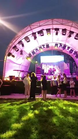 A group of people stand on a grassy area in front of a stage set up for an outdoor event. The stage is covered by a large, arched canopy illuminated with purple and white lights. Several spotlights are attached to the canopy, and a large screen displays a bright image on the stage. On the stage, musical equipment including keyboards, drums, and speakers are visible. Among the people in front of the stage, some are conversing while others appear to be watching the stage. The sky is dark, indicating it is evening or night.