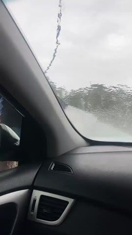 The video begins from the perspective of someone inside a car, looking through the windshield. Raindrops are visible on the glass, distorting the view of the outside. The interior of the car, including a dashboard vent and part of the side mirror, is in focus. As the video progresses, a man appears outside the car, on the left side of the frame. He is holding a blue hose connected to something off-camera. He is wearing a dark shirt. He brings the hose towards the windshield of the car, appearing to spray water or a cleaning solution onto it, as indicated by the water streams on the glass. The man's face is visible, and he appears to be focused on the task. The background outside the car shows trees and buildings, suggesting an outdoor location, possibly a car wash or a service station, given the presence of a large blue and yellow sign with text that is partially obscured. The man continues to operate the hose, cleaning the windshield.