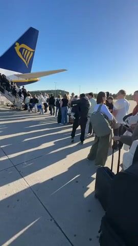 The video captures an active outdoor scene at an airport in Weeze, Germany, under a bright, clear sky.

A large blue and white Ryanair Boeing 737 aircraft dominates the left side of the frame. The airline's distinctive harp logo is prominent on the vertical stabilizer, which also bears the "RYANAIR" name in yellow lettering. The aircraft registration "EI-IGI" is visible near the tail section on the fuselage. The engine beneath the wing also has "RYANAIR" printed on it.

A steady stream of passengers, comprising adults of various ages, are engaged in the boarding process. They ascend a set of mobile stairs positioned at the rear door of the aircraft. Many individuals are carrying personal luggage, including backpacks and roll-on suitcases, as they queue on the tarmac, waiting their turn. A few ground crew members are also visible near the aircraft.

The ground is an asphalt tarmac, bathed in bright sunlight. Long, distinct shadows of the people and the aircraft stretch across the concrete, indicating a low sun angle, characteristic of either early morning or late afternoon. A red cart with a hose or cable reel, along with an orange traffic cone, is positioned in the foreground as part of the airport ground equipment. In the background, other airport buildings and infrastructure can be faintly seen. The weather is exceptionally clear and sunny, with an unblemished blue sky overhead, contributing to the strong contrast and bright illumination of the scene.