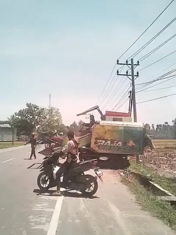 The video depicts a rural roadside scene on a sunny day. A man on a black motorbike waits on the shoulder of a paved road. Behind him, partially on the road and partially on the grassy verge, sits a large, green and yellow combine harvester. A man stands on the road near the front of the harvester, gesturing, while another man is positioned on top of the machine. In the background, a rice paddy extends to the right, while a line of buildings and trees lines the left side of the road. Power lines are visible stretching across the clear sky. A truck passes on the road.