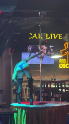 A woman in a blue bikini and black thigh-high boots is performing a pole dance on a small stage with a reflective surface. She’s holding a thin, lighted rod. The stage is surrounded by a gold-fringed skirt. Behind her, a sign reads "CA LIVE" above another sign that says "POOL OSC". There is a screen playing some content. The setting appears to be a bar or club with tables and chairs visible in the background. The lighting is dim with neon accents.