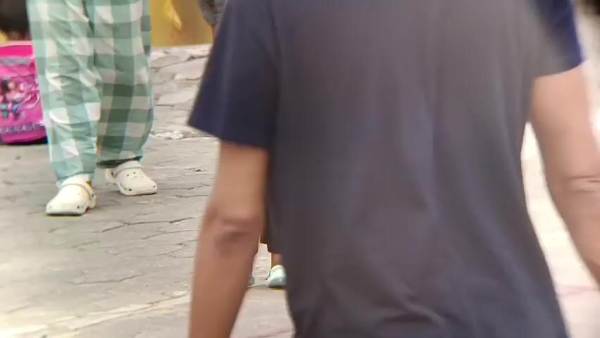 The video shows a young child in a red shirt, black shorts, and a blue cap standing on a paved surface. The child is being hugged by an adult whose arm is visible. To the left, two people are walking away. One is wearing plaid pants and white Crocs, and the other is wearing dark pants. In the background, another person is sitting down and wearing a pink shirt with a cartoon character on it. The scene appears to be outdoors on a street or pathway.