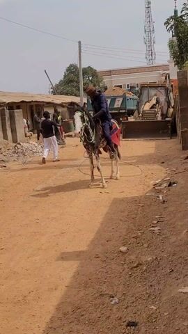 A man is mounting a horse on a dirt road. The horse is mostly white with dark speckles. The man is wearing a long navy blue shirt and pants, and a dark brown hat. He uses the saddle to pull himself up onto the horse. There are other people walking around and a large construction vehicle in the background. The road is littered with debris.