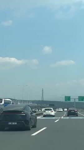 The video shows a view from a car moving on a busy highway under a partly cloudy sky.  Several cars, including a black hatchback in the foreground with the license plate "41 BAT 521," are visible. In the distance, a tall transmission tower is seen. Overhead highway signs display city names, including "Osmangazi," "Körfez," and "Yalova." The road markings are clear and well-maintained. The video is taken during daytime.