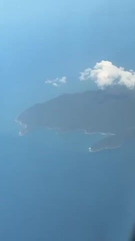 An aerial shot from an airplane shows a large, dark landmass, likely an island or a portion of a coastline, surrounded by a vast expanse of deep blue water. The landmass occupies the upper-middle section of the frame, featuring an irregular, rugged coastline with several coves and points. Above the landmass is a clear blue sky, with a few scattered white cumulus clouds. One larger cloud cluster is positioned slightly above and to the right of the landmass. A very faint, small, white streak is barely visible in the upper-center of the sky. The scene is static, with no discernible movement.