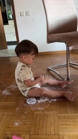 A toddler is sitting on a wooden floor covered in white powder, likely flour or cornstarch. The toddler is wearing a light-colored onesie with an animal print. They are surrounded by the powder, which is also on their legs and hands. A small, round container, possibly for the powder, is next to them on the floor. A pink object, likely a toy, is visible in the lower right corner of the frame. In the background, a kitchen area with cabinets and appliances can be seen, as well as a chair with a metal frame. The toddler appears to be looking down at their hands.