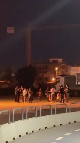 A group of approximately ten people, appearing to be young adults, are gathered in a loosely formed circle in an outdoor area at night. They are illuminated by artificial lighting that casts a warm glow on the ground. In the background, a tall construction crane stands prominently against the dark sky. Behind the crane, several buildings are visible, with some lights on in their windows. A truck is parked to the right of the frame. The people in the circle seem to be interacting with each other, with some holding what appear to be instruments, possibly drums, and engaging in rhythmic activity. The foreground of the image features a concrete barrier with metal railings, suggesting the scene is near a walkway or road. The overall impression is of an informal gathering or performance taking place outdoors after dark.