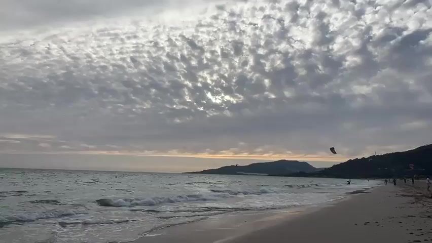 A wide shot of a beach scene at dusk. The sky is overcast with dramatic, textured clouds that obscure the setting sun, though a faint glow of light can be seen on the horizon. Gentle waves lap the sandy shore, creating a foamy white surf line. In the distance, a chain of hills or mountains forms the coastline. Several people are visible on the beach, appearing as small silhouettes walking along the shore. More prominently, there are two individuals kiteboarding on the water. One kiteboarder is closer to the foreground, with their kite clearly visible in the air above them. Another kiteboarder is further out in the water. The overall mood is serene and coastal, with a hint of activity from the water sports.