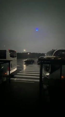 The video shows a dark, rainy outdoor scene. In the foreground, two buses are parked on either side of a pedestrian crossing. The pedestrian crossing is illuminated by white stripes on the wet asphalt. In the background, a black car is driving across the pedestrian crossing. A small blue light is visible in the sky, possibly a drone or a distant aircraft. The overall atmosphere is gloomy and somewhat mysterious due to the darkness and rain.
