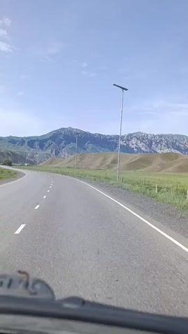 The video shows a first-person perspective of driving on a paved road with a white dashed line on the left and a solid white line on the right. The road curves gently to the left. On the right side of the road, there are solar-powered streetlights. The landscape is characterized by rolling hills covered in green grass, with larger, rugged mountains in the background. The sky is blue with scattered white clouds. The lower portion of the video shows the dashboard of the vehicle.
