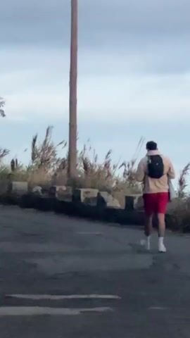 A man with a backpack is running up a paved road. The man is wearing red shorts, white socks, and white running shoes. He is also wearing a light-colored jacket and has a towel slung over his shoulders. A telephone pole is beside the road. Weeds are along the road. The sky is overcast.