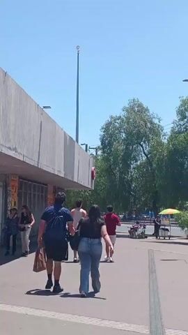 The video shows a paved public plaza on a bright, sunny day.  Several people are walking on the pavement, including a couple walking with a shopping bag.  A row of mature trees borders the plaza. Along the left side is a long, gray concrete building with a blank facade above a lower band of retail spaces with closed, metal security doors. A tall light pole stands between the building and the trees.  In the distance are other people and a vendor pushing a food cart. The sky is clear and blue.