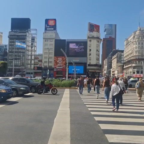 The video shows a daytime street scene in a bustling urban area. Pedestrians are actively using a zebra crossing, moving across the road. Cars are parked along the street, and a motorcyclist is positioned near the curb, seemingly waiting. In the background, tall buildings dominate the skyline, adorned with various billboards and signs. An Argentinian flag is prominently displayed on one of the structures. The sky is clear and blue, suggesting a bright, sunny day.