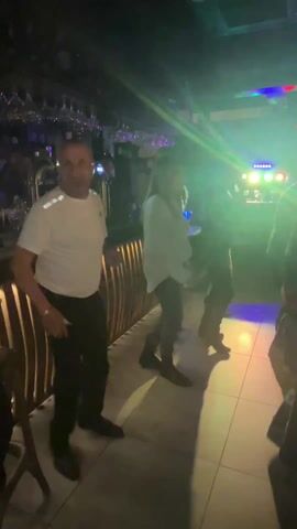 The video shows a group of people dancing at a bar or club. A man in a white t-shirt and black pants is standing near a bar counter, smiling and watching the others. The other people are dancing with the lights flashing blue, pink, and green. The bar counter is decorated with vertical wooden slats. Wine glasses hang from the ceiling. The floor is tiled.
