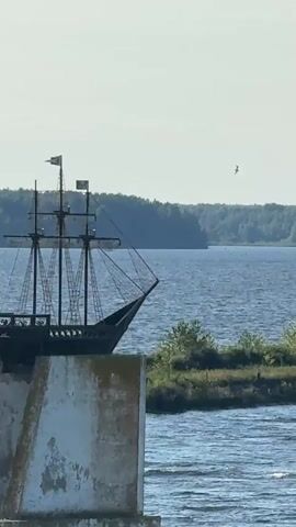 A static, eye-level medium shot captures a black, miniature replica of a three-masted sailing ship mounted on a white, weathered concrete structure. The ship has two flags on its highest mast. The background features a wide expanse of blue water, with a distant treeline of green deciduous trees under a pale sky. Small green bushes and grass are visible on a narrow strip of land to the right of the concrete structure. The water is choppy, indicating some wind.