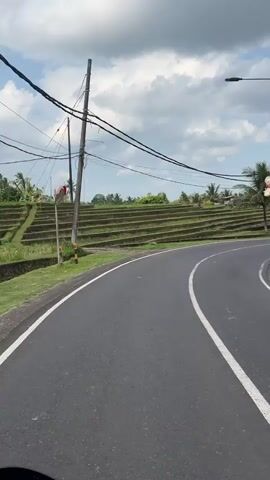 A point-of-view shot from a vehicle moving along a paved road curves to the right. To the left of the road are terraced rice paddies with some patches of green grass. Palm trees and other vegetation are visible. A utility pole with numerous wires is on the left side of the road, near the edge of the rice paddies. A convex mirror is attached to the pole. Further up the road, there are some houses and other vehicles. The sky is partly cloudy with white and gray clouds against a blue background. The road has a white line marking the lane. The bottom of the video shows a portion of the vehicle's dashboard.