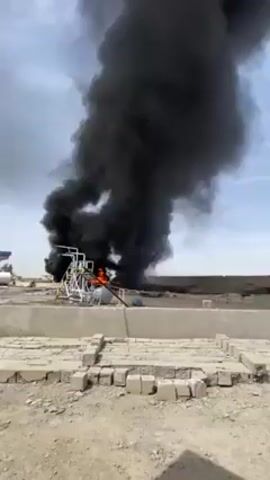 The video shows a large fire with thick black smoke billowing into the sky. The fire is centered around a structure with metal piping and what appears to be a tank. The structure is on a concrete slab surrounded by loose soil and debris. The sky is partly cloudy, and in the background, there's a low-lying building or structure. The fire is substantial, with visible flames consuming the structure.